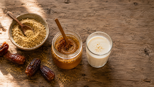 Bowl of golden date palm pollen powder with raw honey being stirred, a glass of warm milk and fresh dates on a wooden table