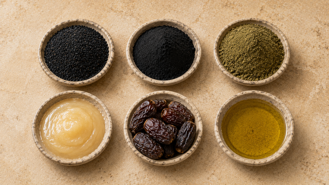 Six dishes arranged in a grid holding black seeds, fine black mineral powder, leaf powder, creamy honey, dates and olive oil on warm cream stone