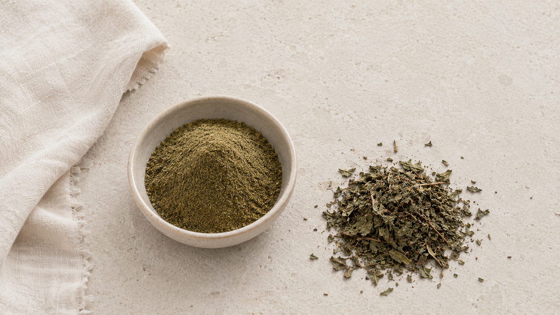 Plain ceramic bowl of green-brown botanical powder beside the dried leaves it was milled from and folded undyed linen on pale stone