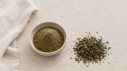 Plain ceramic bowl of green-brown botanical powder beside the dried leaves it was milled from and folded undyed linen on pale stone