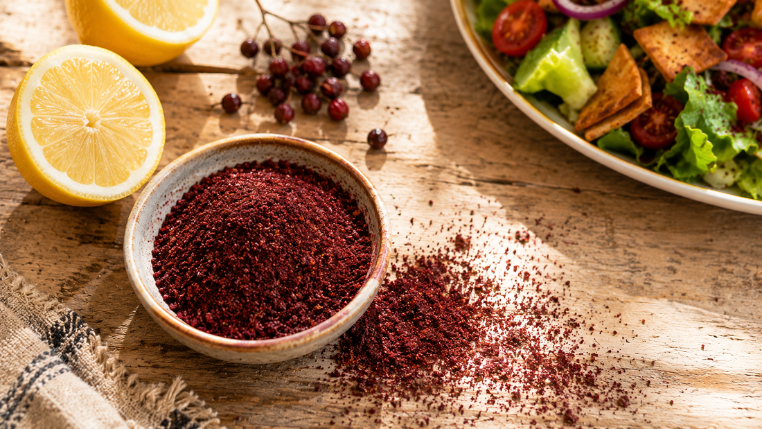 Bowl of deep crimson coarse sumac powder scattered on rustic wood with dried sumac berries and fattoush salad behind