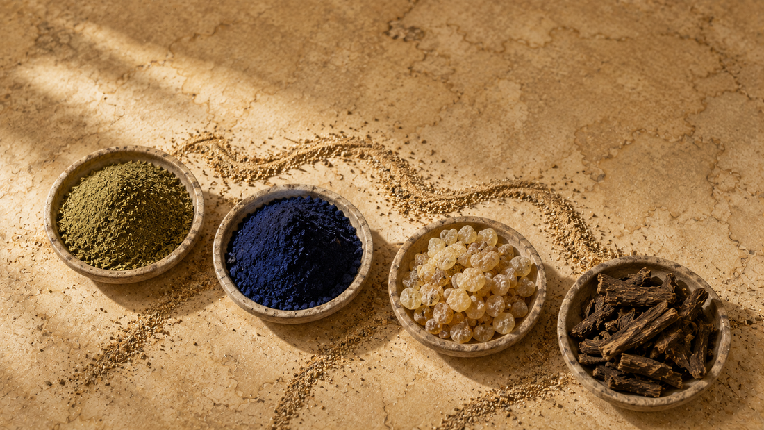 Four dishes of regional botanicals — green leaf powder, indigo blue powder, golden resin and dried root — arranged on a parchment-toned surface with scattered sand