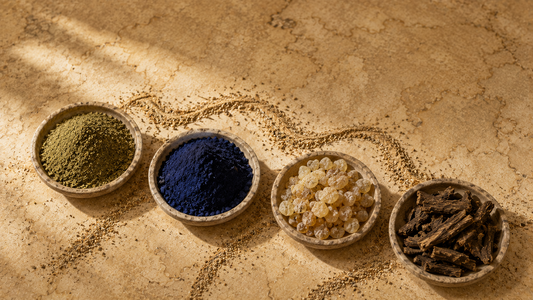 Four dishes of regional botanicals — green leaf powder, indigo blue powder, golden resin and dried root — arranged on a parchment-toned surface with scattered sand