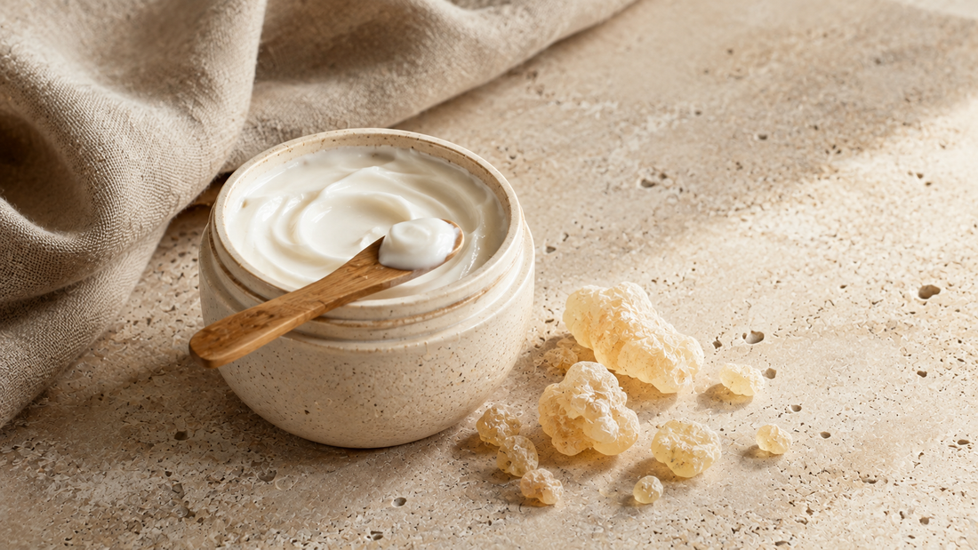 Open ceramic jar of frankincense cream with a wooden spatula, pale frankincense resin tears and folded linen on warm stone