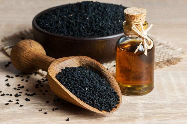 Nigella seeds in a wooden scoop, bowl, and glass bottle of oil on wooden table